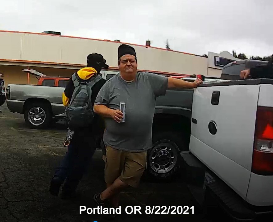 Screenshot of Eddie Orton leaning on a white pickup truck, holding a beer in his right hand, in Portland OR on august 22nd, 2021. 